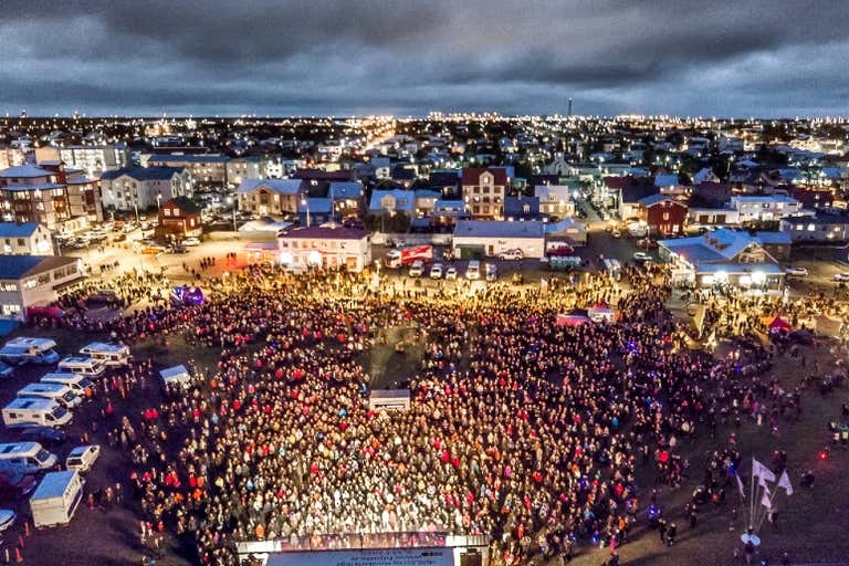 Una gran multitud se reúne en la plaza de una ciudad al atardecer con las luces urbanas de fondo.