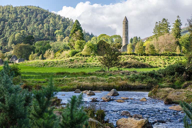 A scenic view of Glendalough with a river, trees, and a round tower.