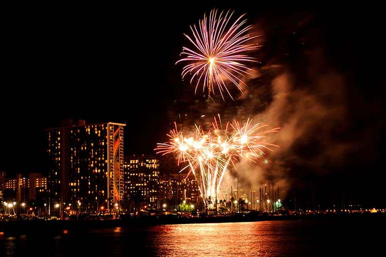 Fireworks lighting up the sky over Honolulu at night, with city lights reflecting on the water.
