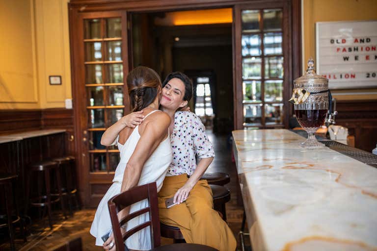 Duas mulheres se abraçam em um bar aconchegante com decoração de madeira e balcão de mármore.