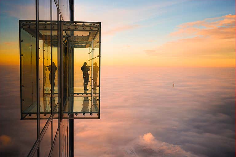 Una persona de pie en una caja de cristal que se extiende desde la Torre Willis sobre las nubes durante la puesta del sol.