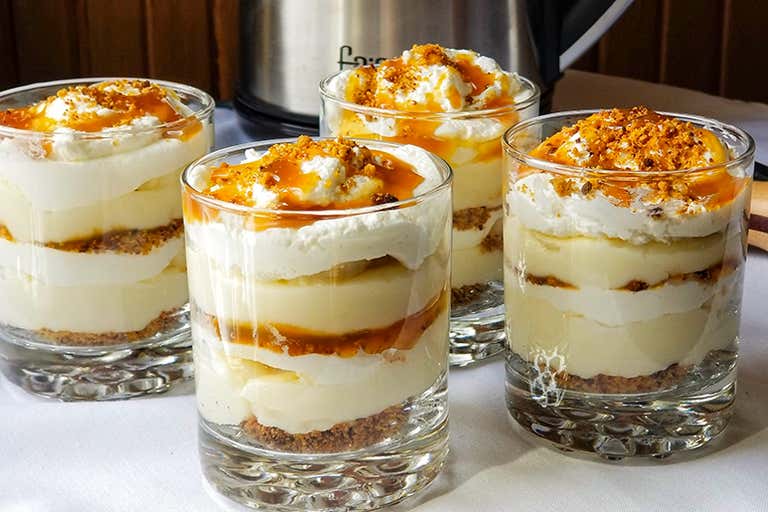 Layered dessert cups with whipped cream, caramel drizzle, and crumbled topping on a white tablecloth.