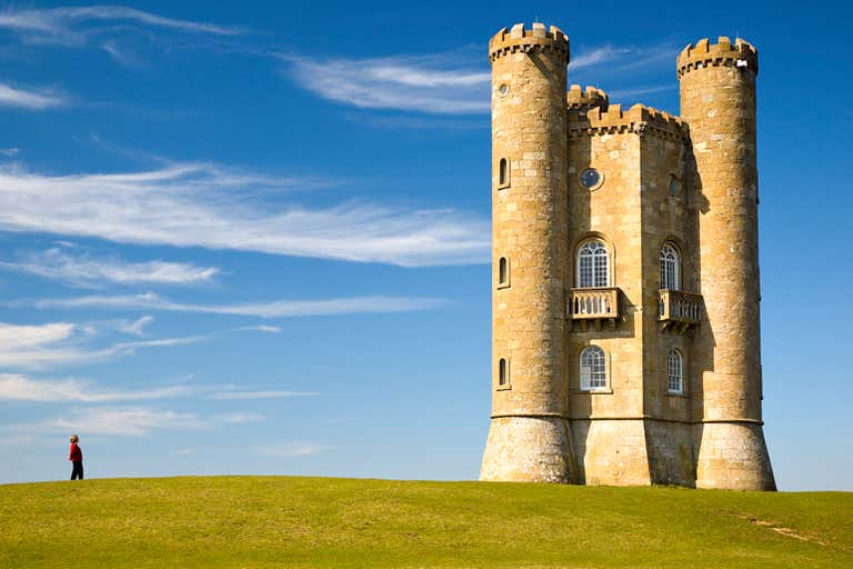 A person walks near Broadway Tower under a clear blue sky.