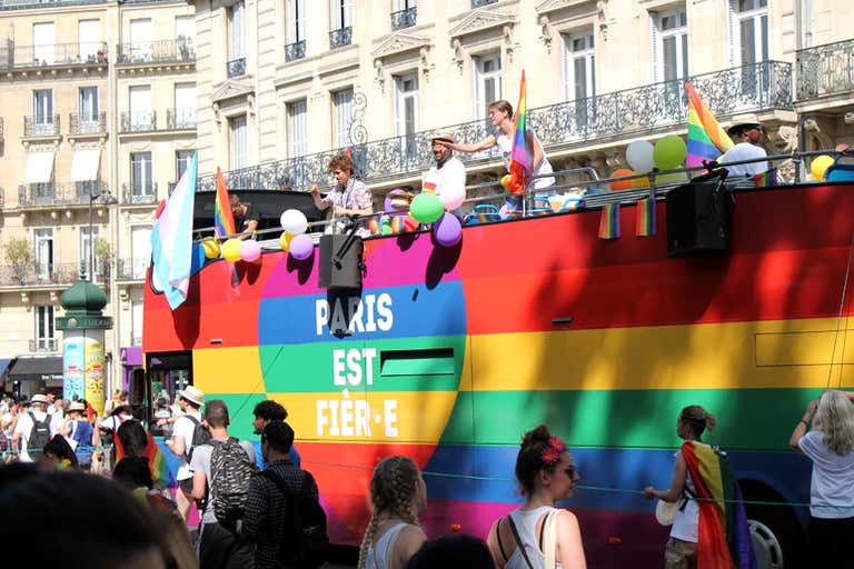 Um vibrante ônibus arco-íris com "Paris est fière" durante a Parada do Orgulho em Paris, com pessoas agitando bandeiras e curtindo a atmosfera.