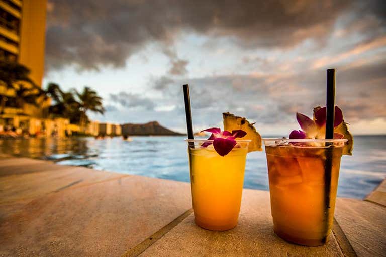 Two tropical cocktails by a pool with Diamond Head visible in Honolulu at dusk.