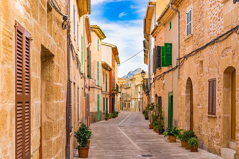 A picturesque narrow street in Alcúdia, Mallorca, with colorful buildings and potted plants lining the pathway.