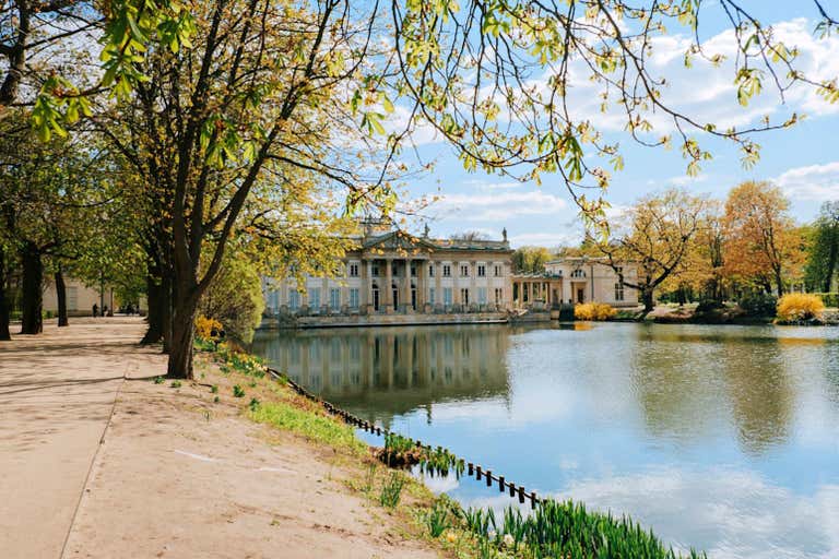 Vista do Parque Łazienki com árvores, um lago e o Palácio da Água.