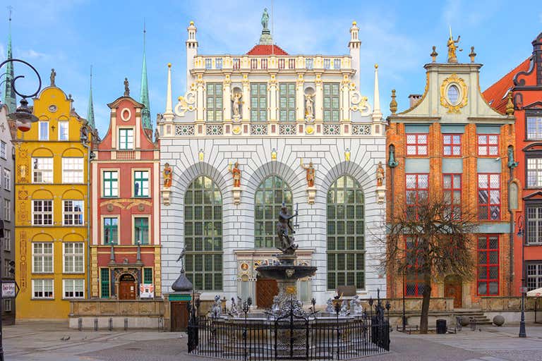 Dwór Artusa w Gdańsku z okazałą fasadą, fontanną Neptuna przed budynkiem i otoczony kolorowymi kamienicami.