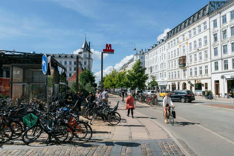 A bustling Copenhagen street with bicycles and pedestrians, surrounded by historic white buildings under a sunny sky.