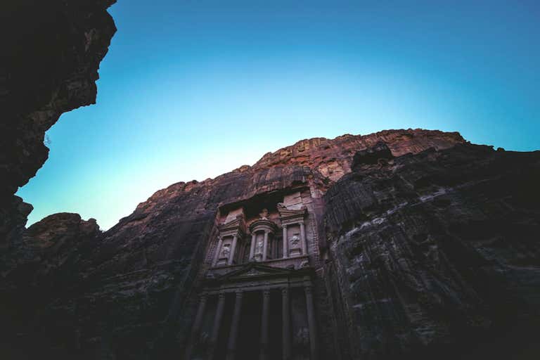 Ancient Treasury building carved into red rock cliff face at Petra, Jordan, viewed from below against bright blue sky.