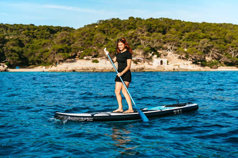 A woman paddleboarding on clear blue waters with a forested coastline in the background.