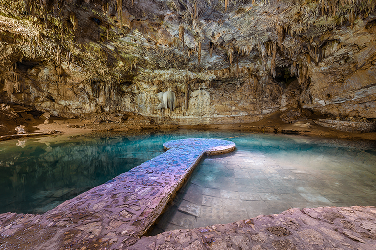 A serene underground cenote with turquoise water, surrounded by stalactites and a stone walkway leading to a platform.