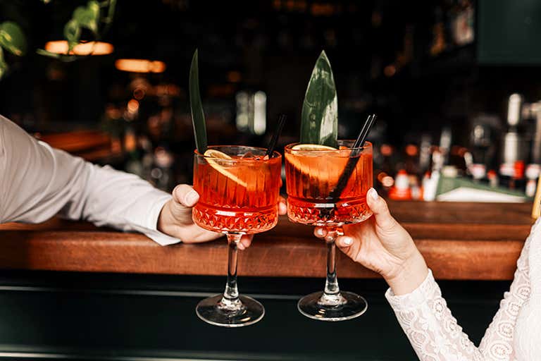 Two people clinking glasses with orange cocktails at a bar.