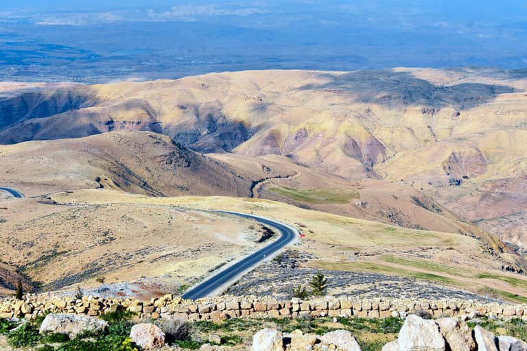 Winding road through barren hills with distant mountains and rocky terrain, viewed from high elevation.