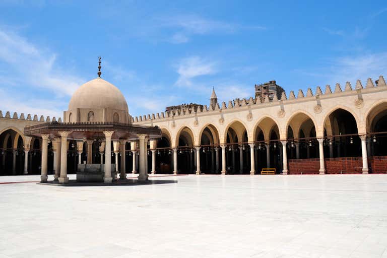The courtyard of the historic Amr ibn al-As Mosque in Cairo, featuring a central dome and arched colonnades under a blue sky.