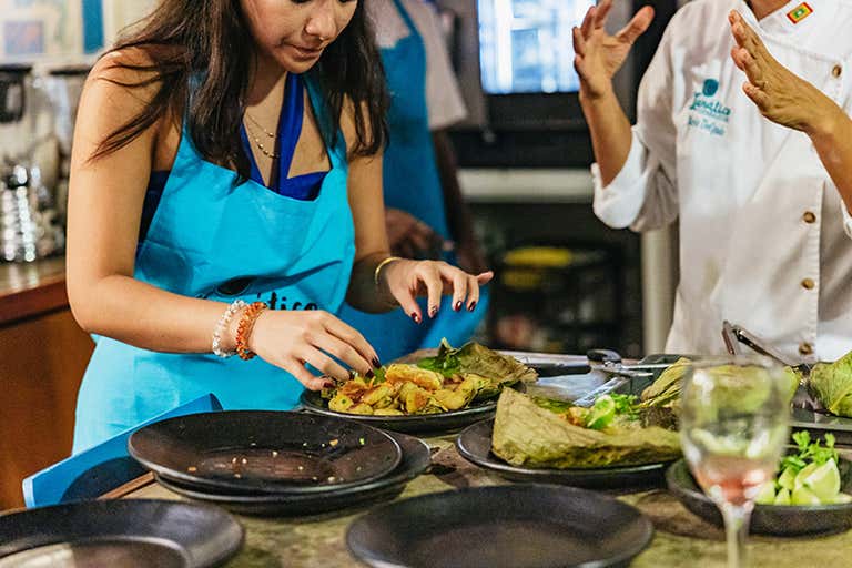 A woman in a blue apron prepares a dish with a chef in a kitchen.