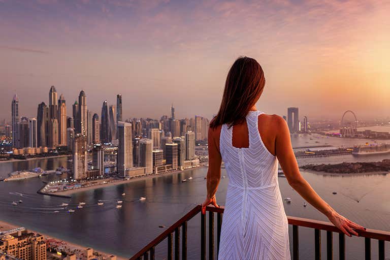 Een vrouw in een witte jurk staat op een balkon dat uitkijkt over Dubai Marina bij zonsondergang.