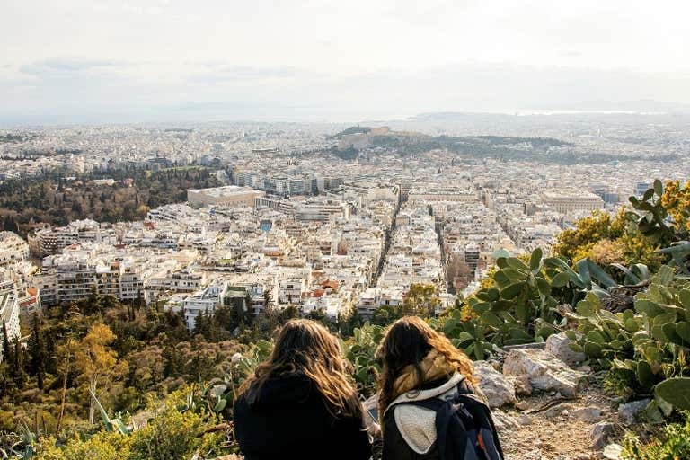 Two people sitting on a hill overlooking the city of Athens with a view of the Acropolis in the distance.