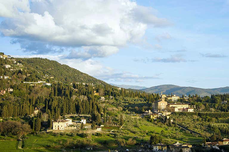 A scenic view of the Tuscan countryside with rolling hills and scattered buildings under a partly cloudy sky.
