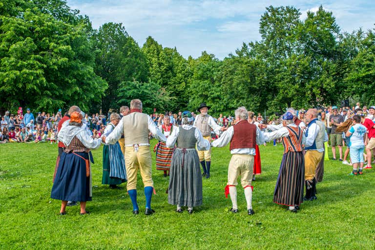 Um grupo de pessoas com roupas tradicionais suecas dançando em círculo em um campo gramado com uma multidão assistindo.