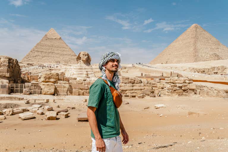 A man standing in front of the Pyramids and Sphinx in Giza under a clear sky, surrounded by sandy terrain.