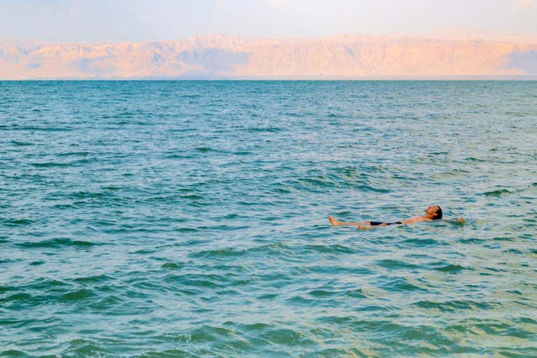 Person floating in turquoise waters of the Dead Sea with distant mountains bathed in sunset light.