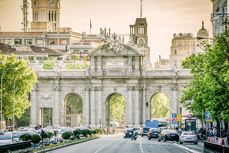 Vista da Puerta de Alcalá, em Madri, com carros e árvores ao longo da rua.