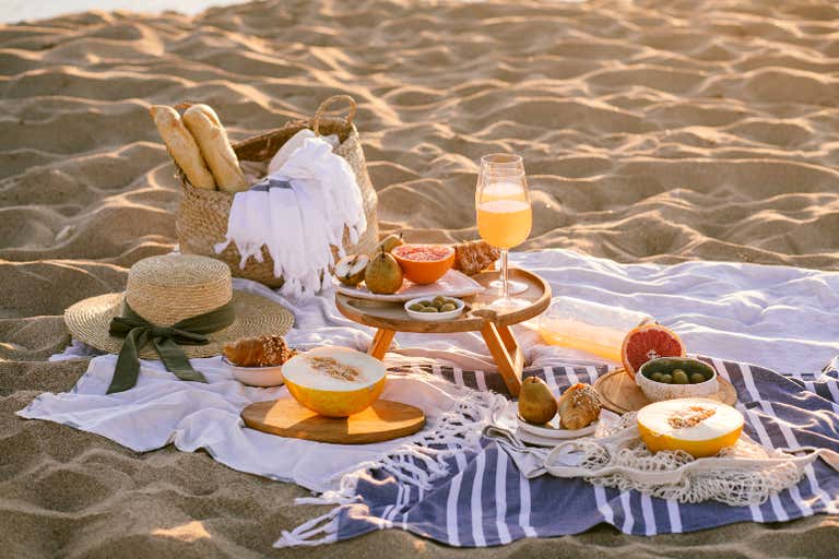 Ein Strandpicknick mit Obst, Brot und einem Getränk im Sand.