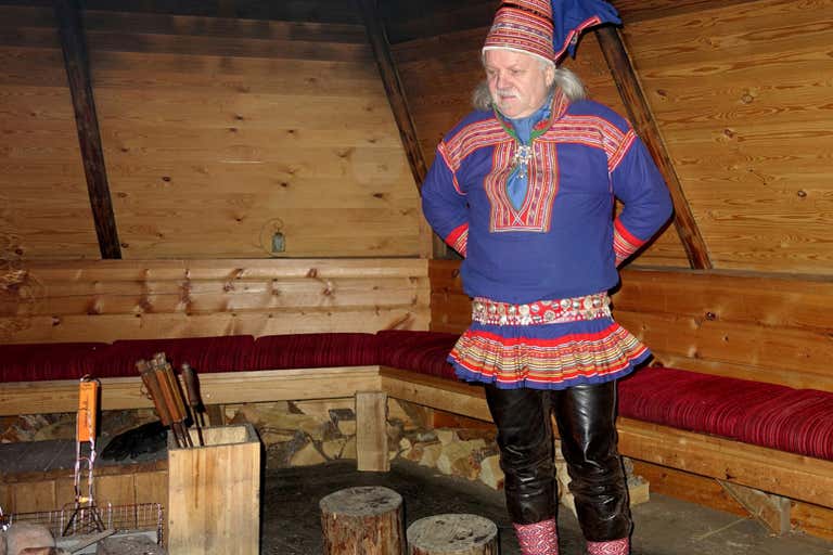 Homem com traje tradicional Sami bordado em azul e vermelho dentro de uma cabana de madeira com lareira.