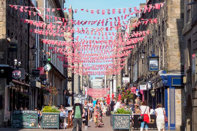 A bustling street in Edinburgh with colorful flags hanging above and people walking along the shops.