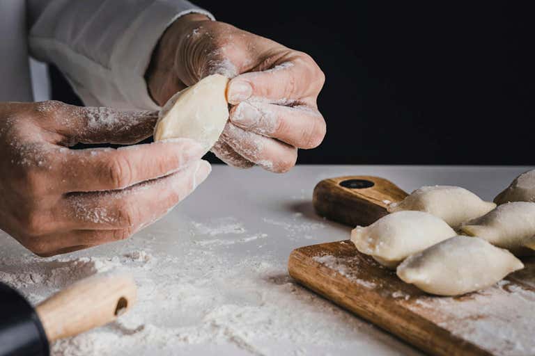 Um conjunto de mãos preparando pierogi fresco em uma mesa com farinha e um rolo de massa.