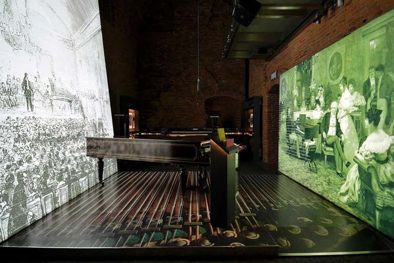 Exposição no Museu Fryderyk Chopin com um piano e projeções históricas nas paredes.
