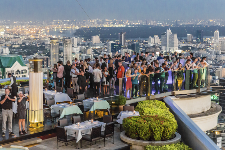 Бар на крыше в Бангкоке, где толпа людей наслаждается видом на город в сумерках.
