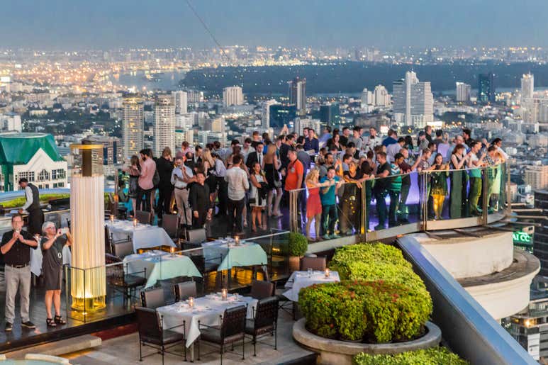Бар на крыше в Бангкоке, где толпа людей наслаждается видом на город в сумерках.