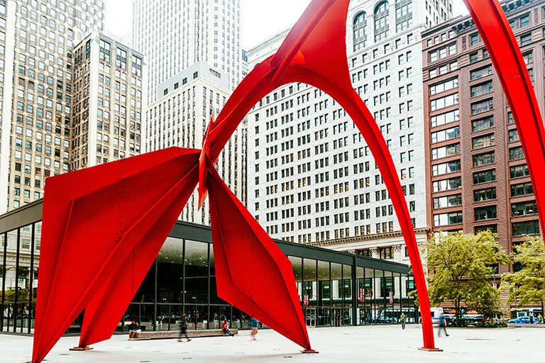 Una gran escultura metálica roja brillante al aire libre, con formas abstractas curvas, se alza en una plaza rodeada de altos rascacielos de Chicago.