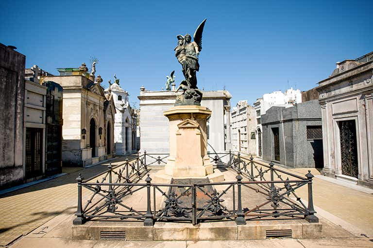 Uma estátua no Cemitério da Recoleta, cercada por mausoléus sob um céu azul claro.