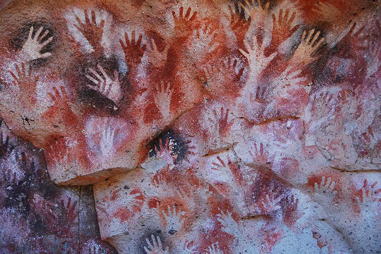 Parede da caverna com várias marcas de mãos em pigmentos vermelhos e brancos.