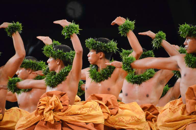 Traditional Hawaiian hula dancers wearing orange skirts and green lei wristbands during a luau in Honolulu at night.