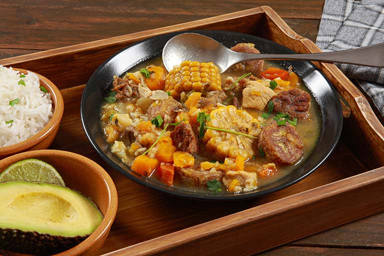 A bowl of sancocho with corn, meat, and vegetables, served with rice and avocado.