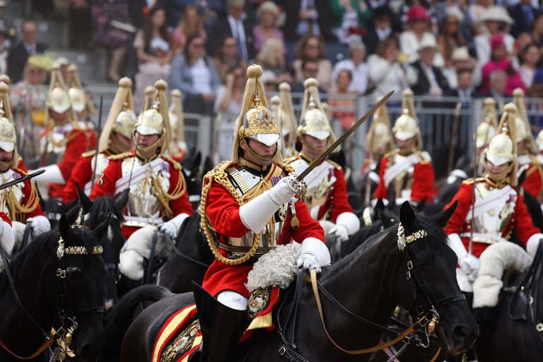 A ceremonial parade with soldiers in red uniforms on horseback, holding swords, in front of a crowd.