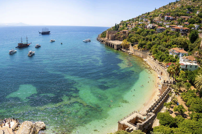 Turquoise Mediterranean waters and beach in Alanya, Türkiye with historic fortress walls, hillside buildings, and tour boats.