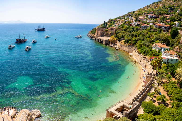Turquoise Mediterranean waters and beach in Alanya, Türkiye with historic fortress walls, hillside buildings, and tour boats.