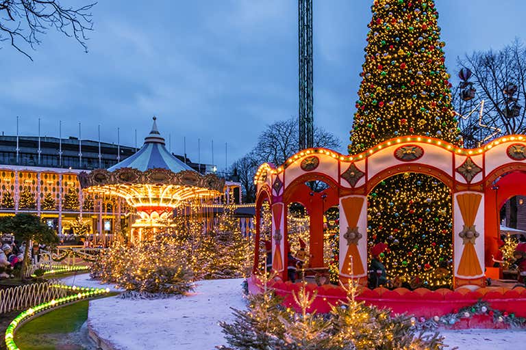 A festive Christmas market with a carousel and lights surrounding a towering Christmas tree at dusk.