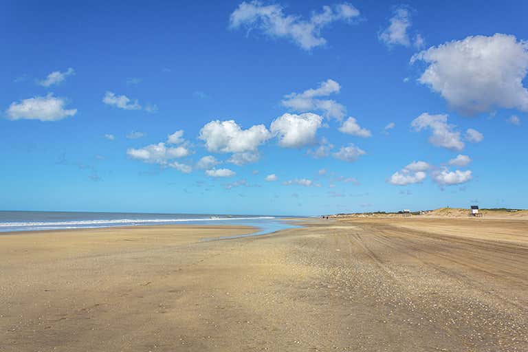 Uma ampla praia de areia com um céu azul claro e nuvens dispersas ao fundo.