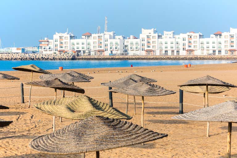 Sombrillas de paja en la playa de arena del Sector Turístico, con los edificios del paseo marítimo al fondo.