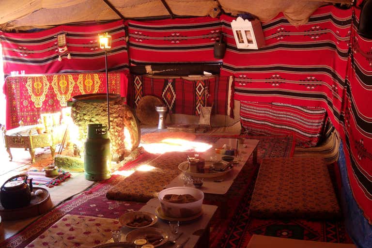 Interior of traditional Bedouin tent with colorful striped textiles, cushions, and low tables set for dining.