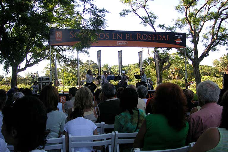 Outdoor Christmas concert in Buenos Aires with audience seated under trees watching performers on stage.