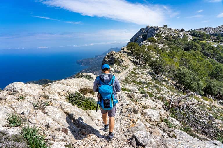 A hiker with a blue backpack walks along a rocky path with a view of the sea and mountains in Mallorca.