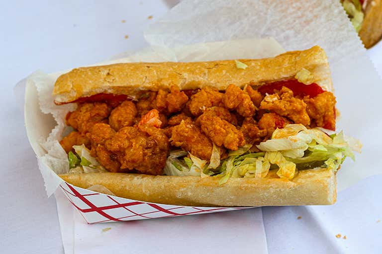 Crispy fried shrimp Po' Boy sandwich with lettuce, pickles, and sauce, served in a paper tray on a white tablecloth.