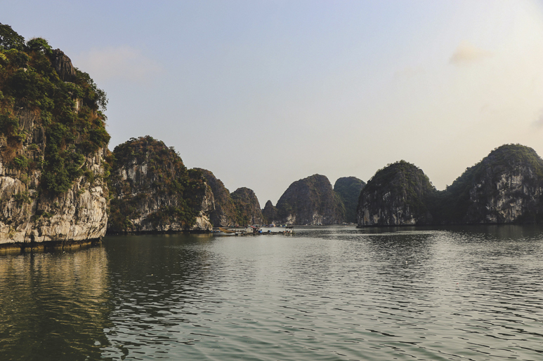 Green-covered limestone karsts of Ha Long Bay stand against a soft sky, and small boats navigating the peaceful waters below.